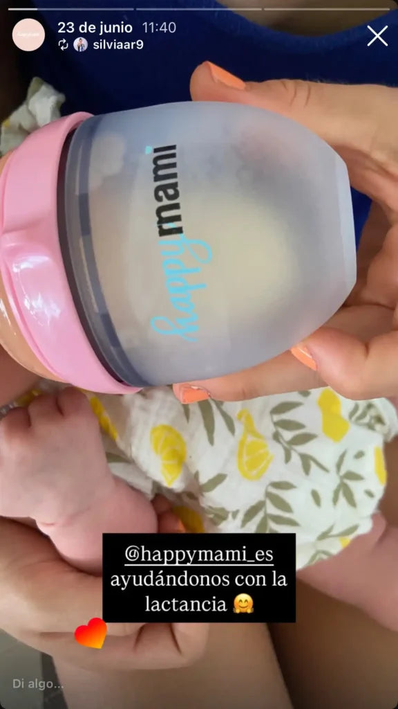 Manos sosteniendo un biberón Happymami rosa con leche junto a un bebé